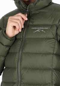Olivgrüne Steppjacke mit durchgehendem Reißverschluss, elastischen Bündchen und Kapuze. Mit dem "SWEDEMOUNT"-Logo in Hellgrau. Strukturierte, gesteppte Optik.