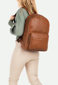 Mochila de cuero marrón con una textura suave, forma redondeada, bolsillo frontal con cremallera y correas ajustables, llevada por una persona.