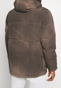 Brązowa kurtka puchowa z kapturem, o lekko błyszczącej fakturze, poziomym pikowaniu i luźnym kroju. Widoczne zagniecenia i ślady zużycia.