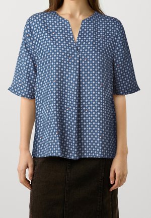 Vrouw draagt een blauw patroonblouse met korte mouwen en een V-vormige inkeping in de halslijn, gecombineerd met een donkerbruine rok, staand tegen een effen achtergrond.