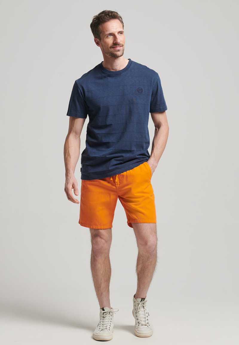 Granatowa koszulka typu crewneck z subtelnymi paskami, w połączeniu z jasnopomarańczowymi szortami. Model nosi białe tenisówki, ręce w kieszeniach, swobodny wygląd.