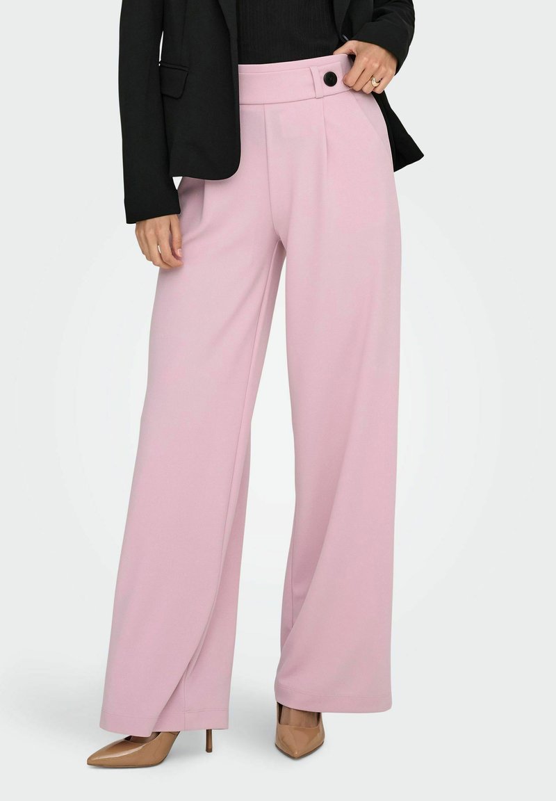 Pantaloni a gamba larga rosa chiaro con una texture liscia, dotati di chiusura anteriore con bottone e pieghe, abbinati a un blazer nero e tacchi beige.