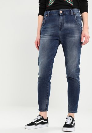 Persoon die versleten blauwe spijkerbroek en zwart-witte sneakers draagt, staand tegen een effen lichte achtergrond.