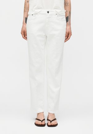 Άτομο με λευκό τζιν με ίσια μπατζάκια, λευκό μπλουζάκι και μαύρες σαγιονάρες, στέκεται με τα χέρια χαλαρά στα πλευρά.