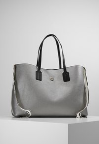 Grand sac cabas argenté avec poignées noires et une bandoulière rayée, posé sur un socle blanc devant un fond uni.
