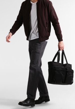 Veste bomber bordeaux foncé, t-shirt blanc, chinos gris, bottines noires à la cheville, et un grand sac fourre-tout noir avec plusieurs compartiments.
