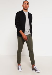 Giacca bomber nera, t-shirt grigia, joggers verde oliva e sneakers grigie. Tessuti lisci e design aderente con polsini e caviglie elasticizzati.