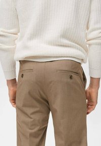 Beige tailored trousers with two back pockets and button accents, worn with a textured cream sweater. Smooth fabric with a fitted design.