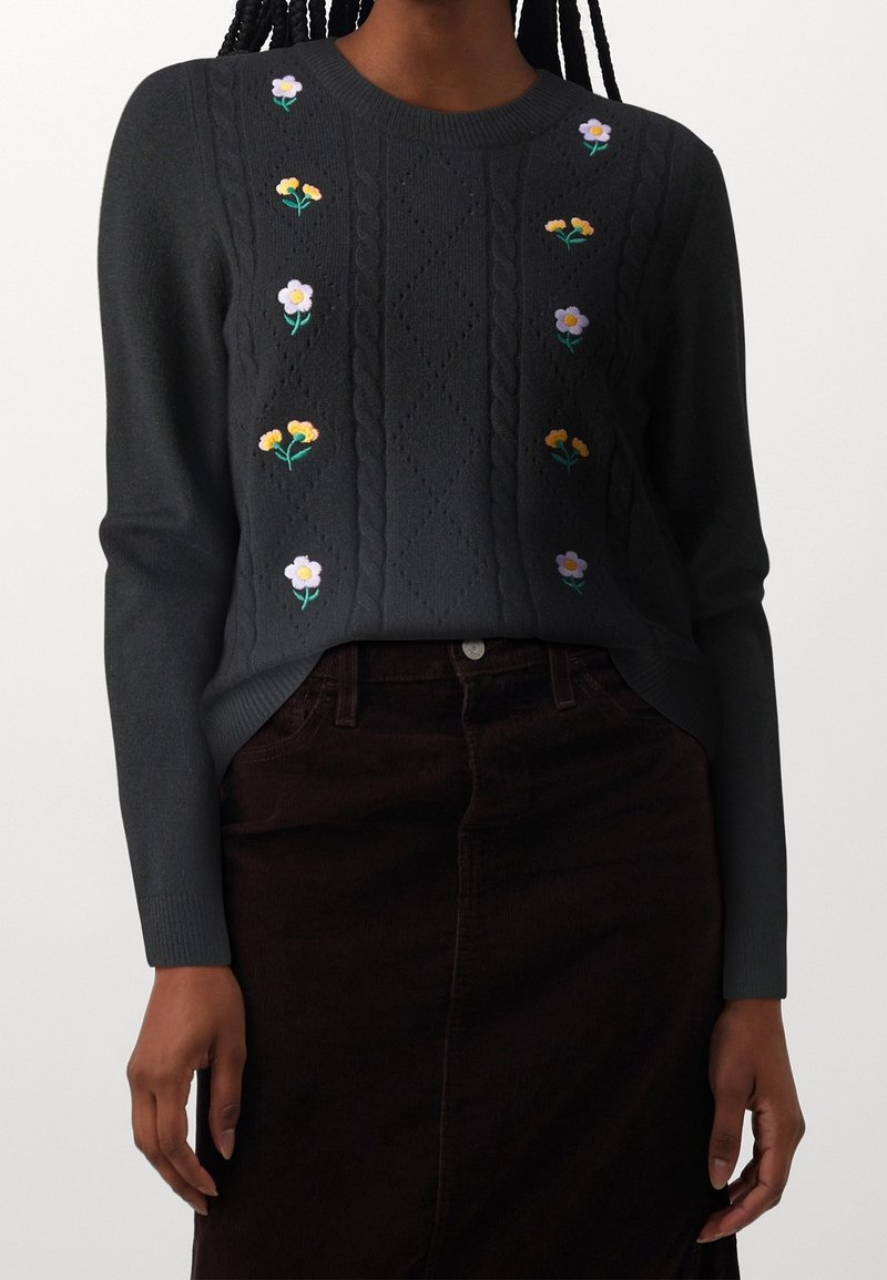 Pull en maille gris foncé avec motifs torsadés et petites fleurs jaunes et blanches brodées, porté avec une jupe sombre.