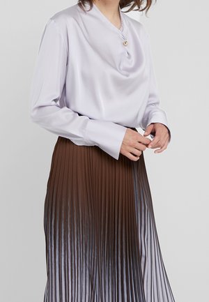 Kobieta ubrana w jasnofioletową satynową bluzkę z perłowym guzikiem oraz brązową plisowaną spódnicę midi, stojąca na jednolitym tle.
