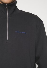 Sudadera negra con media cremallera hecha de tela suave. Cuenta con un pequeño logo azul "TIGER OF SWEDEN" en el pecho y un cuello estructurado.