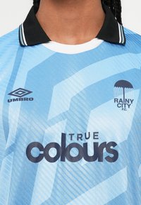 Šviesiai mėlyna futbolo marškinėliai su juodu apykakle, Umbro logotipas, Rainy City F.C. emblemą, kuriame pavaizduota skėčio, ir "True Colours" rėmėjo tekstas.
