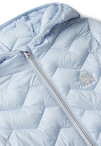 Světle modrá prošívaná bunda se stříbrným zipem a malým vyšitým logem na hrudi, složená tak, aby ukázala kapuci a švy.