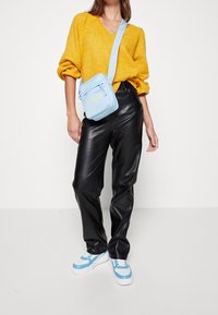 Femme portant un pull jaune moutarde, un pantalon en cuir noir, un sac bandoulière bleu clair et des baskets blanches avec des accents bleus.