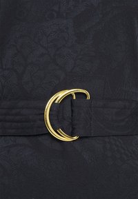 Schwarzer Stoffgürtel mit einem strukturierten Muster, versehen mit einer runden goldenen Metallschnalle. Das Material hat einen subtilen Glanz, der die Designdetails unterstreicht.