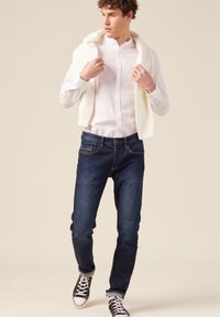 Outfit uomo composto da una camicia bianca con bottoni, jeans scuri in denim blu e sneaker nere con accenti bianchi; maglione appoggiato sulle spalle.