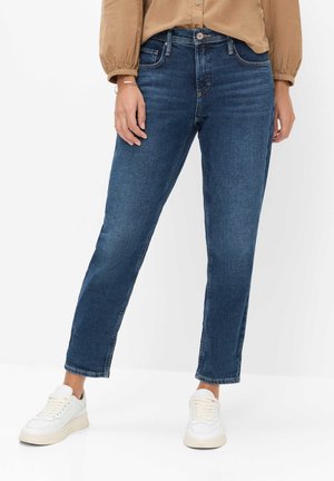 Person trägt blaue Jeans mit geradem Bein, beige langärmeliges Shirt und weiße Sneakers, steht vor weißem Hintergrund.