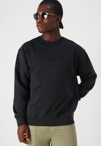 Svart sweatshirt med rund hals och präglad Levi's-logotyp på framsidan. Modellen bär mörka solglasögon, tillsammans med ljusa byxor.