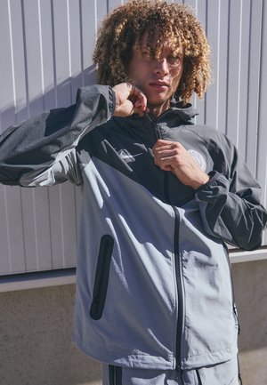Lightweight zip-up jacket in gray and black with a logo, side pockets, and elastic cuffs for a fitted design. Outdoor style suitable for active wear.