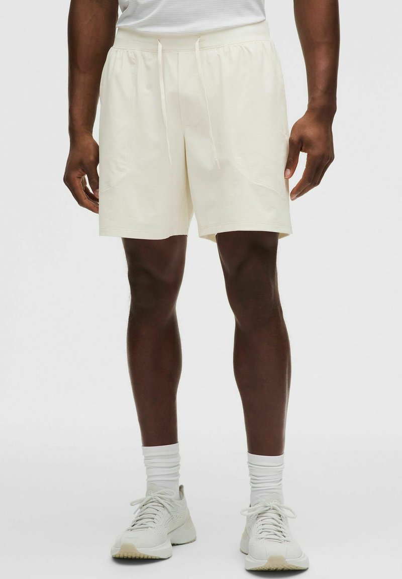 Atleta que lleva puestos pantalones cortos de cuerda color crema, calcetines blancos hasta la mitad de la pantorrilla y zapatos deportivos blancos, de pie sobre un fondo liso.