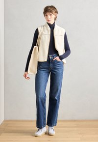 Cream fleece vest with front pockets, navy long-sleeve top, blue straight-leg jeans, and white slip-on shoes. Beige tote bag.