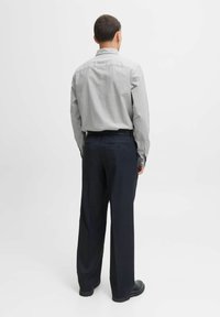 Maglietta grigia a maniche lunghe con polsini a bottone e colletto, abbinata a pantaloni ampi blu scuro. Tessuto dalla texture liscia. Scarpe nere.