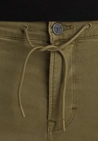 Kaki trenirke s pojasom na vezanje, metalnim gumbom i bočnom džepom. Glatka tekstura s ojačanim šavovima i završecima za vezanje u boji.