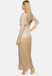 Sukienka maxi w błyszczącym złoto-beżowym gradiencie, z długimi rękawami, dopasowaną talią i gładką teksturą, noszona z beżowymi szpilkami.
