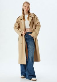 Beige trenchcoat i lättviktsmaterial, med dubbelknäppt design och breda slag, kombinerad med smala blå jeans.