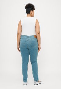 Pantalones vaqueros ajustados en denim azul claro, con un parche de cuero marrón en la parte trasera, cinco bolsillos y una sutil textura desvaída.