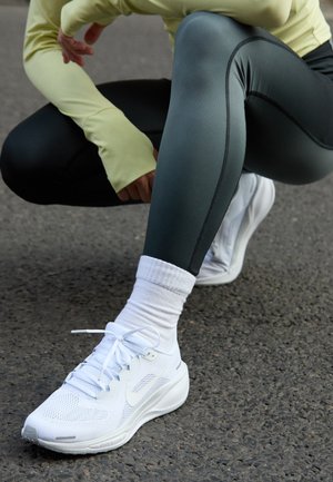 Hvite sportsko med mesh-overdel, flate snørebånd og dempet såle, parret med mørkegrønne leggings og en lys gul langermet topp.