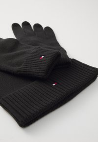 Guantes de punto negro acanalado y gorro a juego dispuestos en plano, cada uno con una pequeña etiqueta rectangular roja, blanca y azul en el puño.