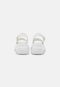 White sandals with a chunky heel and wide straps, featuring an adjustable ankle buckle and a textured rubber sole.