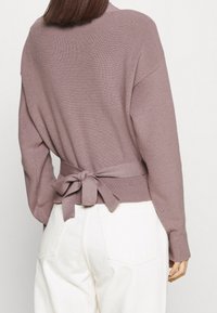 Suéter de punto en malva, con un lazo en la cintura, mangas largas y textura acanalada. Combinado con pantalones blancos de tiro alto.