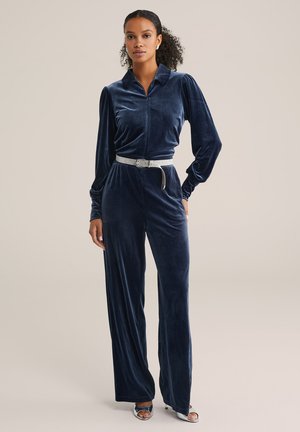 Samtblauer Jumpsuit mit kragenlosem Ausschnitt, langen Ärmeln und einer taillierten Taille, die durch einen silbernen Gürtel akzentuiert wird. Glatte, weiche Textur.