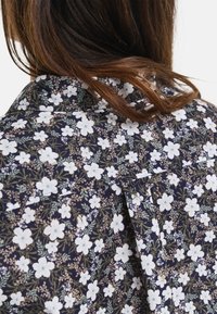 Blusa floreale con sfondo blu navy scuro, decorata con fiori bianchi e foglie verdi. Tessuto morbido con vestibilità rilassata e dettaglio con piega sul retro.