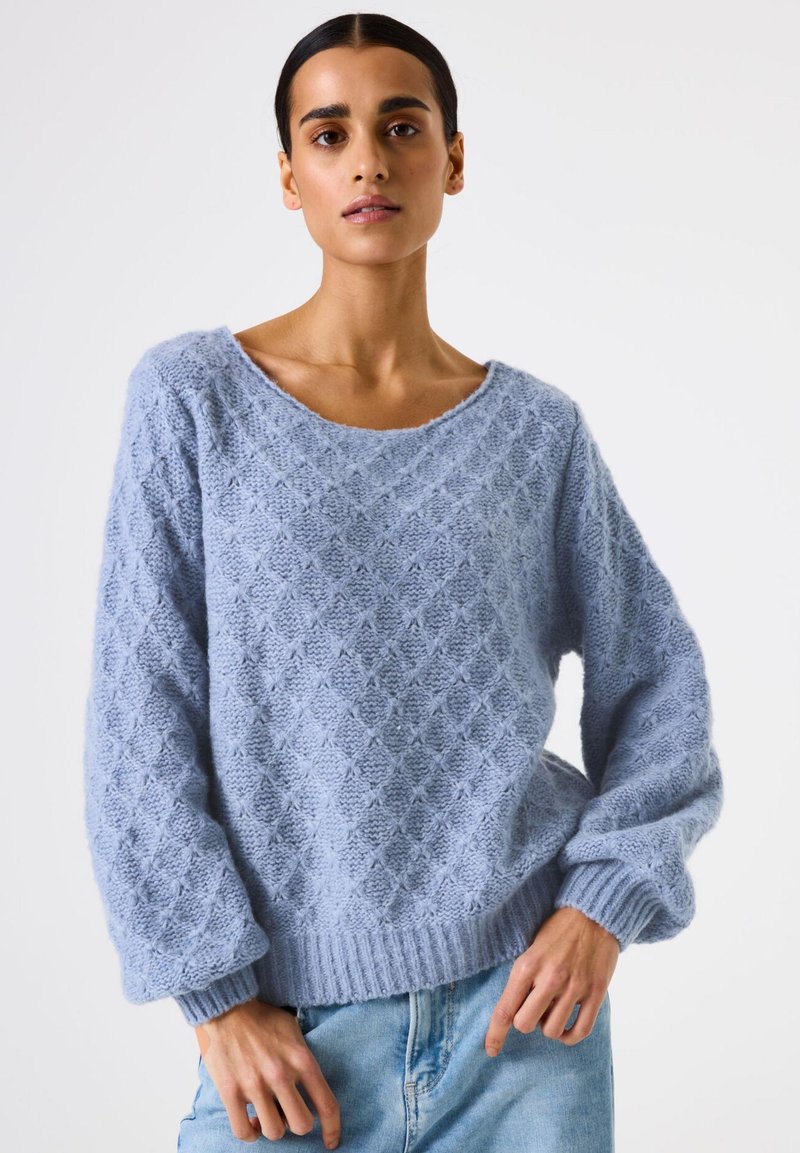 Femme portant un pull en tricot texturé bleu clair avec des manches ballon et un jean bleu clair, se tenant devant un fond blanc uni.