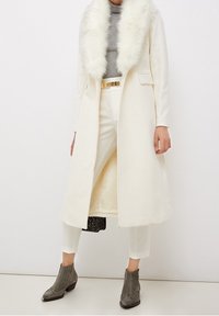 Manteau en laine blanche avec col en fausse fourrure, associé à un haut en maille gris, un pantalon blanc et des bottines grises ornées de franges.