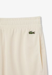 Shorts en maille couleur crème avec une taille élastique et un logo crocodile vert brodé sur le côté. Présente plusieurs petits trous de ventilation.