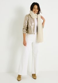 Veste bomber en or métallisé, écharpe crème, jean évasé blanc et bottes à talons couleur sable. La veste possède une fermeture éclair à l'avant et des poignets côtelés.