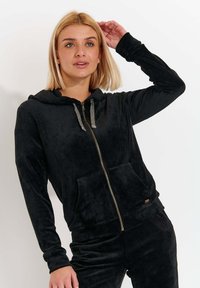 Felpa nera con zip realizzata in morbido tessuto peluche. Presenta un cordino a contrasto, tasche laterali e una vestibilità comoda.