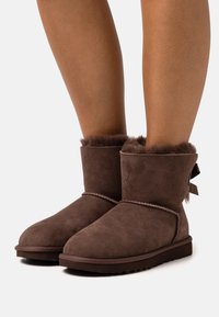 Bruna mocka ankelboots med en mjuk, fluffig foder och en liten rosett på baksidan, med en platt gummisula.