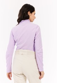 Donna che indossa una giacca in pile viola chiaro e pantaloni beige, rivolta di spalle, con capelli scuri su uno sfondo bianco semplice.