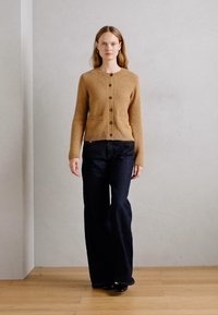 Cardigan in lana beige con chiusura a bottoni, due tasche frontali, indossato sopra dei jeans in denim scuri a zampa. Il modello è in piedi davanti a uno sfondo neutro.
