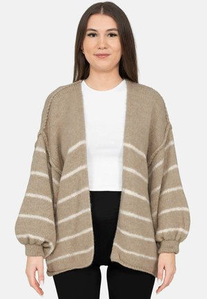 Frau trägt eine beige gestrickte Strickjacke mit weißen horizontalen Streifen, ein weißes Oberteil und schwarze Hosen, steht vor einem schlichten Hintergrund.