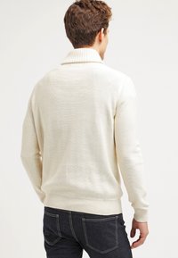 Sweter w kolorze kremowym, z wysokim kołnierzem, ściągaczami na rękawach i u dołu, z fakturowaną powierzchnią. Stylizowany z ciemnymi dżinsami.