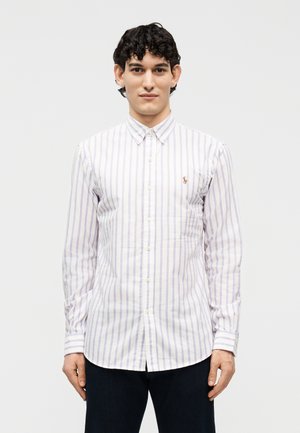 Άνδρας που στέκεται προς τα εμπρός φορώντας λευκό πουκάμισο με κουμπιά και μακριά μανίκια με ανοιχτό μωβ κάθετες ρίγες και σκούρο παντελόνι.