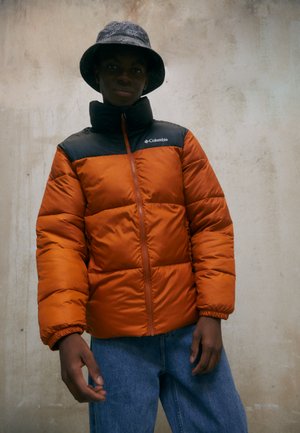 Giacca piumino arancione con colletto nero, dotata di zip frontale e polsini elasticizzati. Abbinata a jeans di denim blu e un cappello a secchiello con texture.