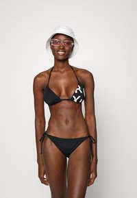 Čierne bikiny s trojuholníkovým vrchom a bielym nápisom, doplnené nízko strihanými spodkami na šnúrky. Model má na sebe biely klobúk a ružové slnečné okuliare.