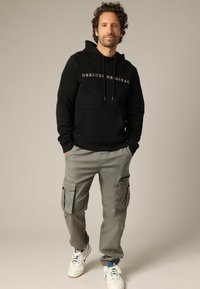 Zwarte hoodie met witte tekst "DEELUXE ORIGINAL", gecombineerd met grijze cargobroek met meerdere zakken en een relaxte pasvorm. Witte sneakers.